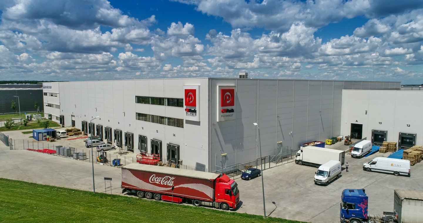 Delivery trucks maneuver at a large white logistics distribution center.